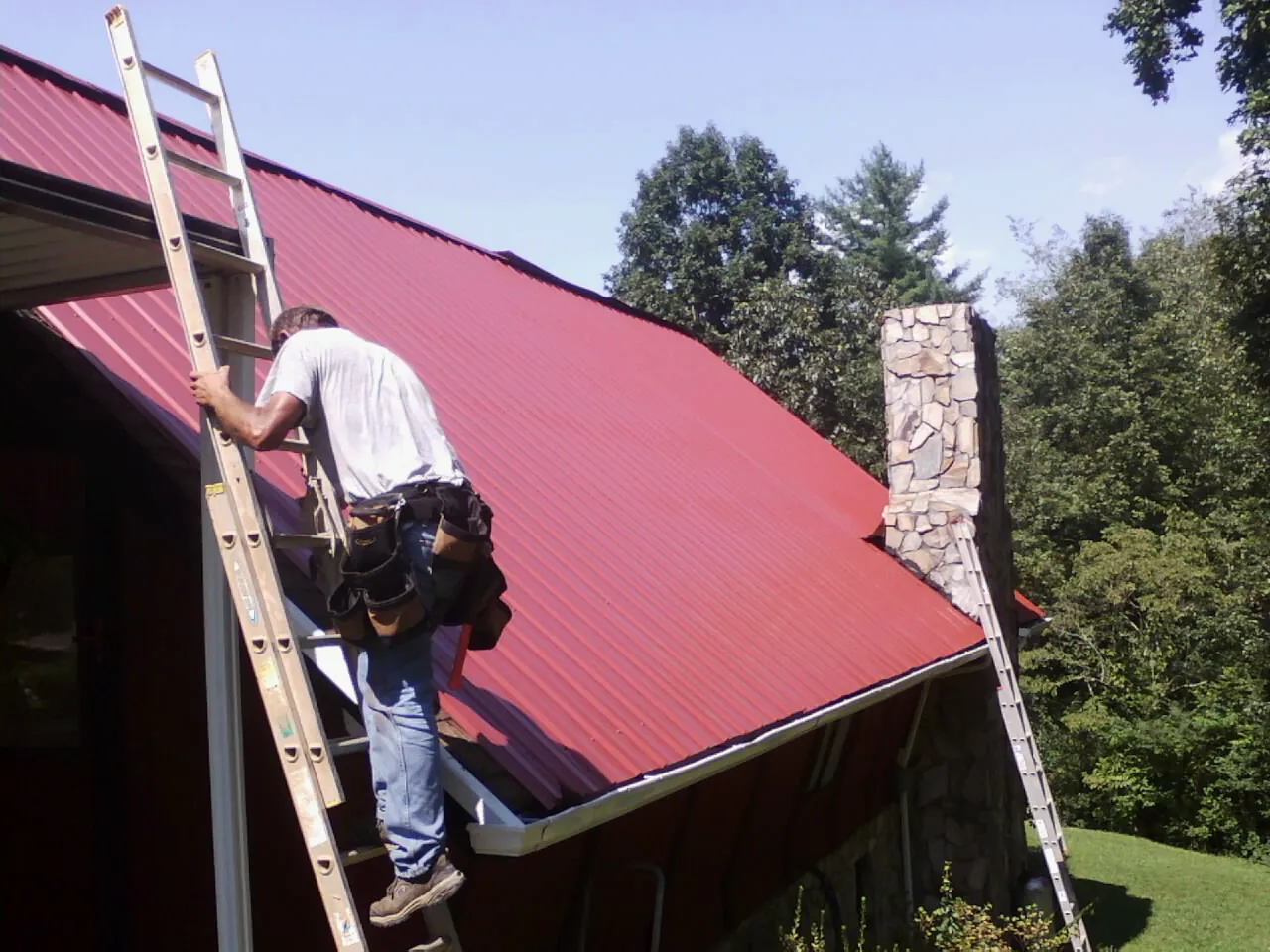 Roofer on a ladder beside a red metal roof and stone chimney — Soffit & Fascia Repair by Roofing Brothers in Winner